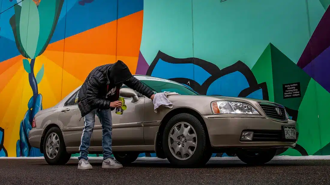 Una persona está limpiando la rueda delantera derecha de un sedán plateado estacionado frente a una pared mural vibrante y multicolor. El mural presenta grandes formas geométricas en tonos de azul, verde, naranja y morado. La persona se inclina sobre la rueda con un paño en la mano, vistiendo una chaqueta negra, jeans azules y zapatillas. La matrícula del vehículo es visible.