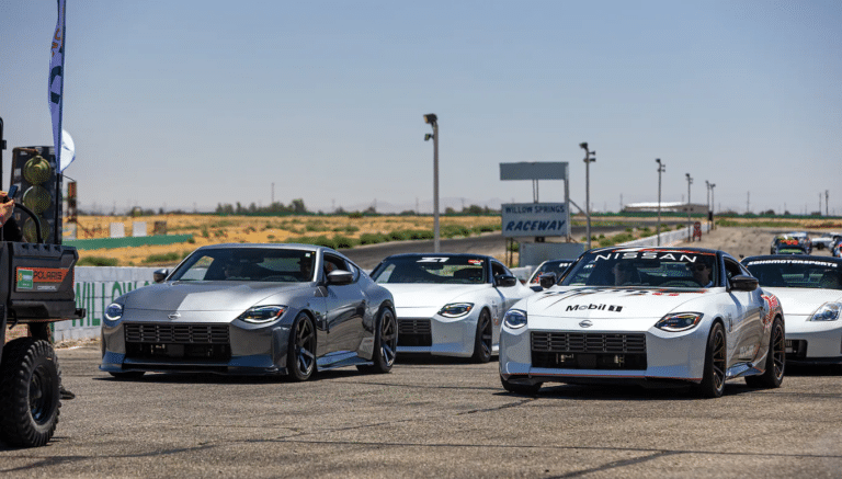 Una flota de coches Nissan Z alineados en una pista de carreras durante el evento ZCON. El coche más cercano está decorado con calcomanías de carreras, incluyendo el logo de Mobil 1, y está seguido por varios otros de color y diseño uniformes. Al fondo, hay un cielo azul claro y un cartel que dice ‘Willow Springs Raceway’, indicando la ubicación del evento.