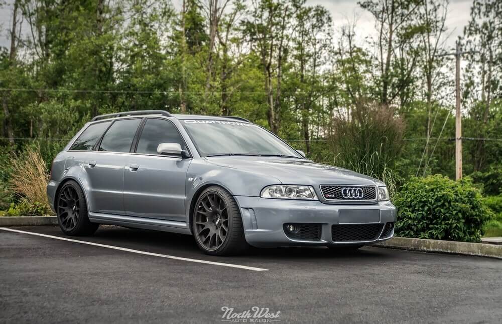"La imagen muestra un Audi plateado estacionado en un aparcamiento. El coche tiene una apariencia deportiva con grandes llantas de aleación negras, ventanas tintadas y una parrilla frontal con el logotipo de Audi y una insignia 'S', indicando que es un modelo de alto rendimiento. El parabrisas tiene una calcomanía que dice 'QUATTRO', refiriéndose al sistema de tracción total de Audi. El fondo consiste en vegetación y árboles, sugiriendo que el coche está estacionado en un entorno natural o suburbano. Esta imagen es interesante porque muestra una versión de alto rendimiento de un vehículo práctico, combinando utilidad con deportividad.
