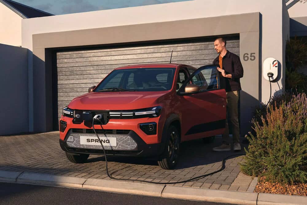 Imagen de una persona junto a un coche eléctrico Dacia Spring de color naranja, estacionado frente a un garaje. El coche está conectado a una estación de carga montada en la pared a la derecha de la puerta del garaje. El número '65' es visible en la pared junto al garaje. La persona parece estar interactuando con el coche, posiblemente abriendo o cerrando la puerta. La escena se desarrolla en una zona residencial, y el coche está posicionado en un camino pavimentado.