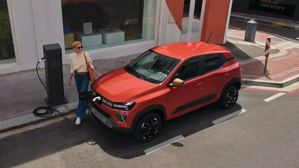 Dacia Spring color naranja-rojo en un punto de recarga eléctrica, fotografiado desde un ángulo elevado. El vehículo está estacionado junto a un poste de carga negro, con una persona conectando el cable de carga. El coche muestra su diseño crossover compacto con detalles en negro como las llantas y las molduras laterales. La escena se desarrolla frente a un escaparate con ventanales grandes y paredes blancas y naranjas. El pavimento muestra marcas de plaza de aparcamiento y hay un poste indicador en la esquina.