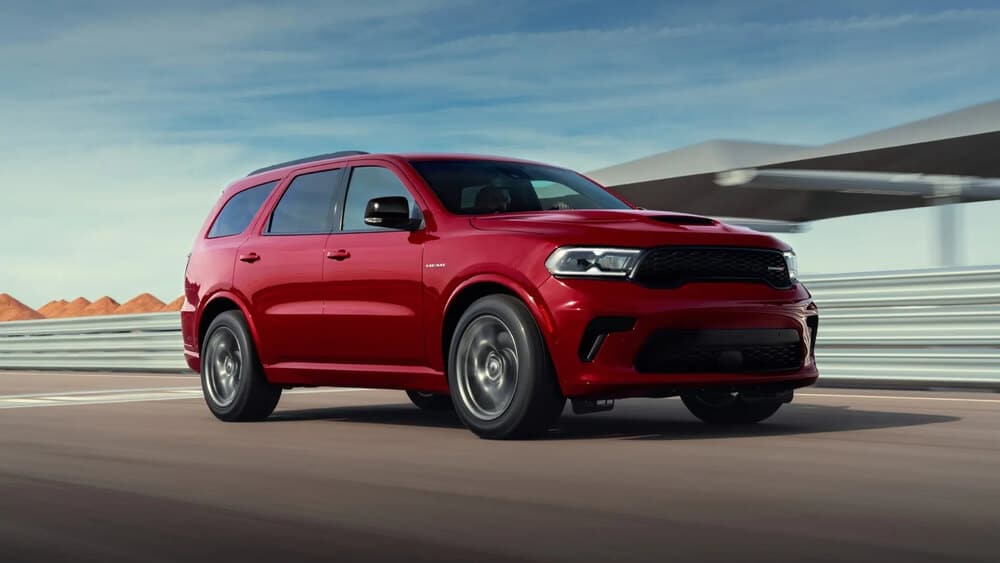 Esta imagen muestra un Dodge Durango de color rojo conduciendo por una carretera o pista. El vehículo es un modelo de alto rendimiento, equipado con un motor HEMI V8, conocido por su potencia y capacidad. El fondo incluye un cielo despejado y algunas estructuras, posiblemente parte de una pista de carreras o una instalación de pruebas. Esta imagen es interesante porque destaca las capacidades dinámicas y el diseño del vehículo, lo que puede atraer a los entusiastas de los automóviles o a posibles compradores que buscan un SUV potente y elegante