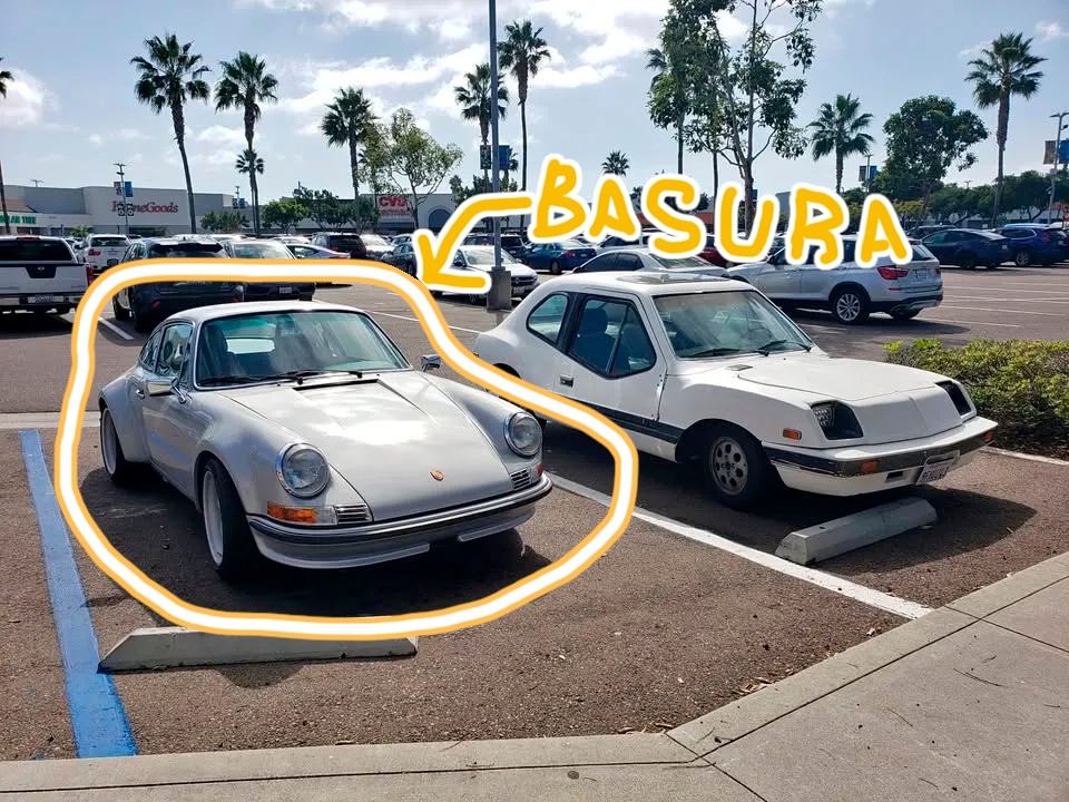 Dos autos estacionados en un estacionamiento al aire libre. El auto a la izquierda es un Porsche clásico plateado, mientras que el auto a la derecha es un Electrek Unancar blanco. Encima del Electrek Unancar hay un texto escrito a mano que dice 'BASURA' con una flecha apuntando hacia el auto. El Porsche está rodeado por un contorno naranja, destacándolo en la imagen. Al fondo, se pueden ver otros autos estacionados y algunas palmeras.