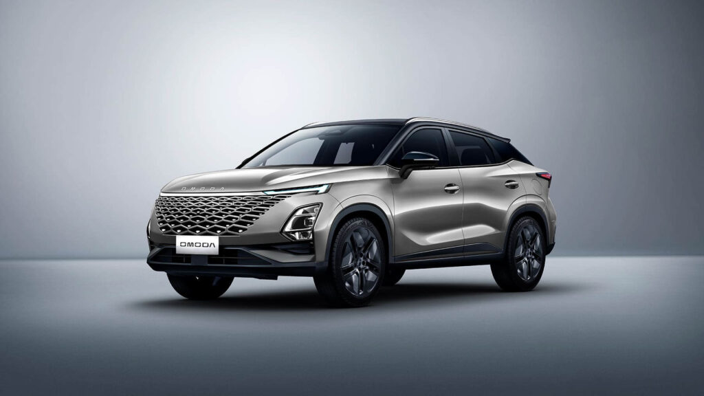 Fotografía promocional del SUV Omoda 5 en color plata metalizado sobre un fondo gris neutro. El vehículo se muestra en ángulo tres cuartos delantero, destacando su diseño moderno con una llamativa parrilla frontal en patrón diamante, faros LED angulares, línea de techo tipo coupé con techo negro contrastante, y llantas de aleación deportivas. La carrocería presenta líneas fluidas y dinámicas con marcados pliegues laterales. El diseño general transmite una imagen premium y deportiva característica de los SUV crossover contemporáneos.