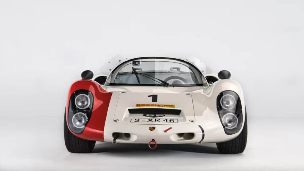 Vista frontal de un automovil de carreras Porsche 910, con el número 1 en el capó. El coche es blanco con detalles en rojo y negro, y tiene faros redondos dobles a cada lado. En el capó se pueden ver varias etiquetas, incluyendo una de Bosch y otra de BP, así como la matrícula S-XR 46.