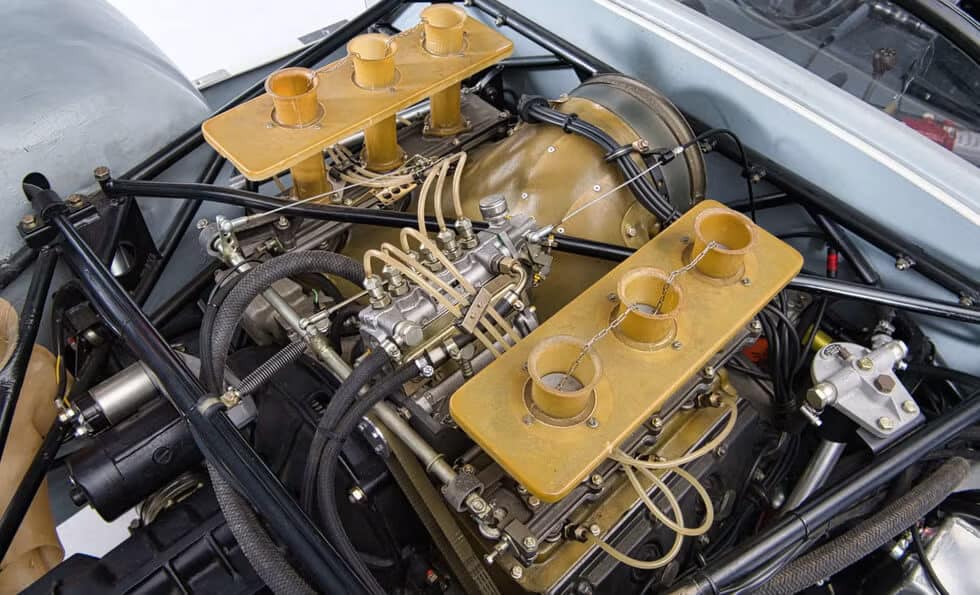 Vista detallada del motor de un Porsche 910, enfocándose en el sistema de admisión. El motor presenta cuerpos de aceleración individuales con pilas de velocidad, diseñados para mejorar la eficiencia y el rendimiento de la admisión de aire. También se puede ver la intrincada red de líneas de combustible y enlaces, indicando una configuración de alto rendimiento, probablemente para un vehículo de carreras o de alto rendimiento.