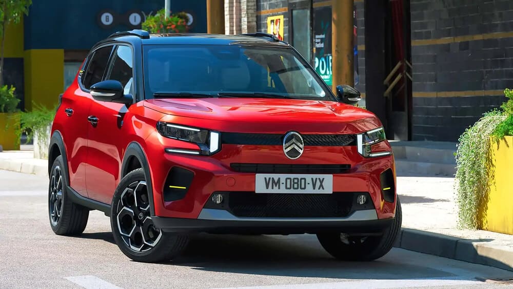 Imagen de un Citroën e-C3 rojo, un SUV compacto eléctrico, estacionado en una calle. El vehículo presenta un diseño moderno con faros LED distintivos, un techo negro y llantas de aleación elegantes. La matrícula dice 'VM-080-VX'. El entorno parece ser urbano, con vegetación y edificios al fondo. Esta imagen es relevante ya que destaca el diseño y las características del Citroën e-C3, mostrando su atractivo en un entorno urbano.