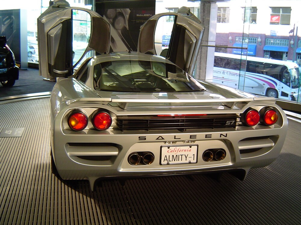 "La imagen muestra la vista trasera de un Saleen S7 plateado con sus distintivas puertas de ala de gaviota abiertas. El coche está exhibido en un interior, probablemente en una sala de exposición, y está posicionado sobre una plataforma circular. La matrícula dice 'ALMITY-1' e indica registro en California. El Saleen S7 es conocido por su alto rendimiento y diseño único, lo que lo convierte en un tema interesante y relevante para los entusiastas de los automóviles. El fondo incluye grandes ventanas que muestran una escena urbana con edificios y un autobús.