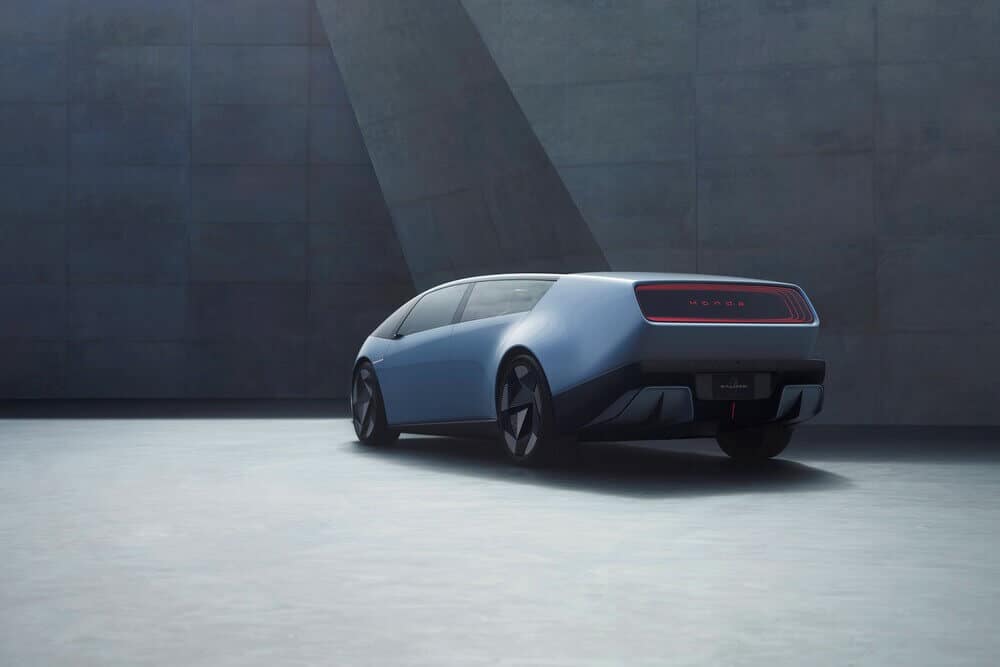 Imagen de un automóvil Honda 0 Saloon de color azul claro con un diseño aerodinámico y ventanas tintadas oscuras. La parte trasera del coche presenta una tira de luz horizontal con la palabra 'Honda' iluminada en rojo. El coche está posicionado en un entorno industrial moderno con un suelo de concreto y una gran pared texturizada en el fondo. El diseño y el entorno sugieren que se trata de un coche conceptual, probablemente mostrando tecnología avanzada y elementos de diseño innovadores. El nombre 'Saloon' es visible en el área de la matrícula, indicando que es un modelo sedán.