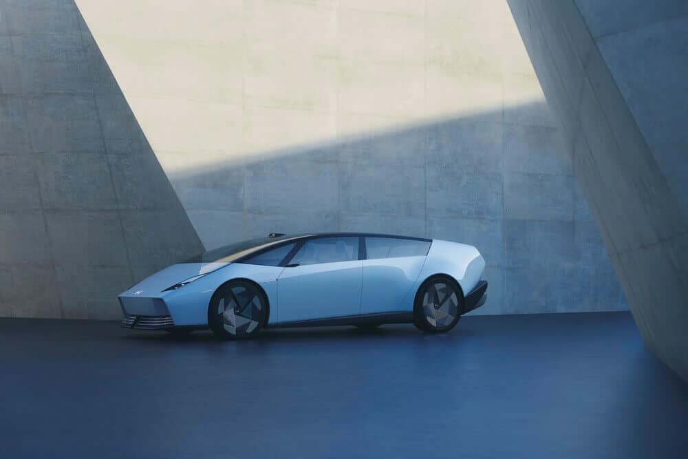 Imagen de un automóvil Honda Series 0 Saloon de color azul claro estacionado en un entorno minimalista con paredes de concreto. El vehículo tiene un diseño aerodinámico y moderno, con líneas limpias y ruedas de aspecto geométrico. La luz del sol crea sombras interesantes en las paredes y el suelo, destacando la forma del automóvil.