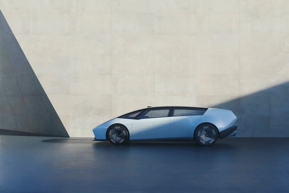 "Imagen de un automóvil Honda 0 Saloon de color blanco visto de perfil, estacionado en un entorno minimalista con paredes de concreto. El coche tiene un diseño aerodinámico y moderno, con líneas limpias y una forma baja y angular que recuerda al Lamborghini Gallardo. Las ruedas tienen un diseño geométrico distintivo y la iluminación crea sombras dramáticas en la pared, destacando la forma del vehículo.