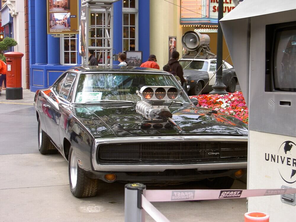 La imagen muestra un muscle car clásico, específicamente un Dodge Charger negro, estacionado en un entorno al aire libre. El coche tiene un supercargador prominente que sobresale del capó, indicando que es un vehículo de alto rendimiento. El entorno parece ser una zona temática, posiblemente un estudio de cine o un parque de atracciones, como sugiere el cartel de Universal Studios y la marca "Fast & Furious" en la cinta de barrera. Hay personas en el fondo, y los alrededores incluyen flores coloridas, un buzón rojo y varias estructuras y señales, añadiendo al ambiente temático.