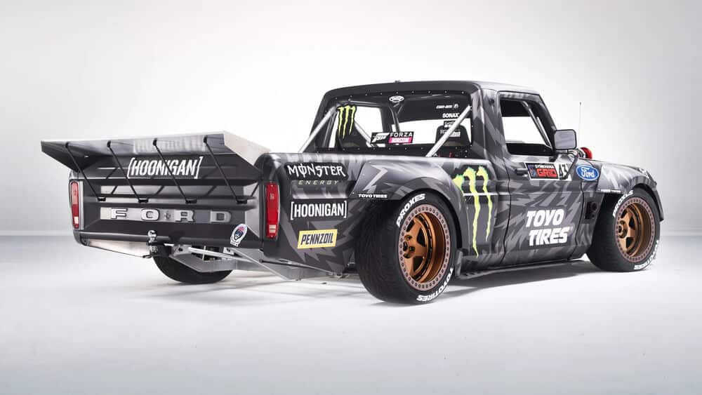 La imagen muestra una camioneta Ford personalizada conocida como el "Hoonitruck". Este vehículo está fuertemente modificado para rendimiento y impacto visual, con un gran alerón trasero, llantas anchas y varios adhesivos de patrocinadores, incluyendo Monster Energy, Hoonigan, Toyo Tires y Pennzoil. La camioneta tiene un distintivo patrón de camuflaje en negro y gris con acentos verdes. Las modificaciones sugieren que está diseñada para actividades de alto rendimiento como drifting o carreras, lo que la hace interesante y relevante para los entusiastas del automovilismo y los fanáticos de los deportes de motor.