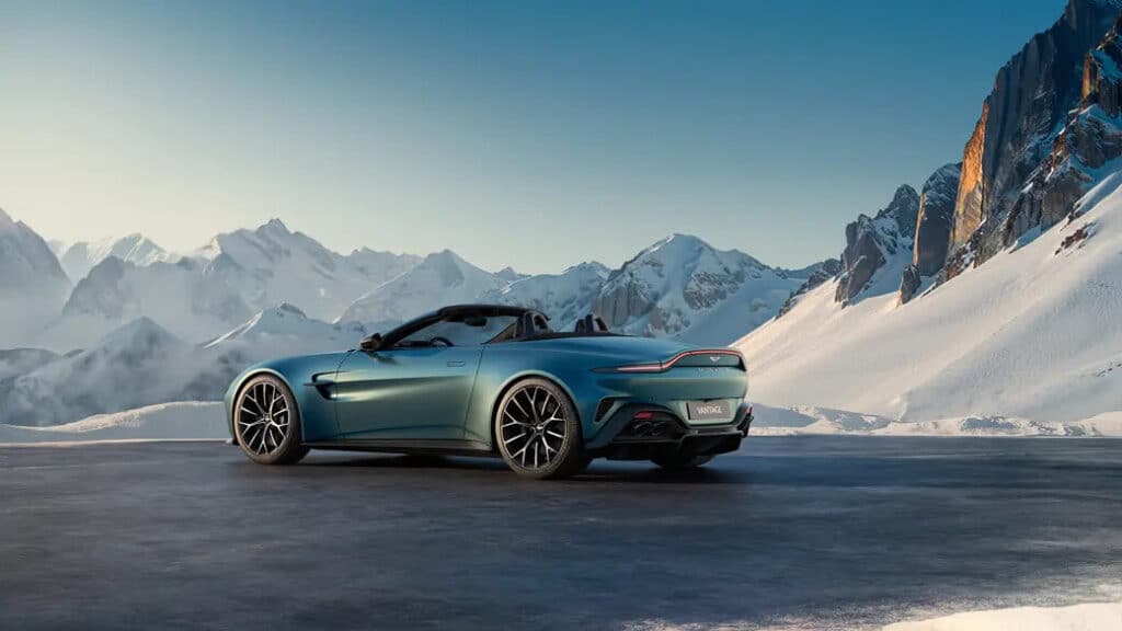 Imagen de un coche deportivo descapotable azul conduciendo por una carretera de montaña con picos nevados al fondo. El coche se desplaza por una carretera con una barrera de piedra al lado, y la escena se desarrolla en un paisaje majestuoso de montañas cubiertas de nieve bajo un cielo despejado. La imagen es interesante y relevante porque captura el contraste entre el diseño moderno y elegante del coche y la belleza natural y robusta del paisaje montañoso.