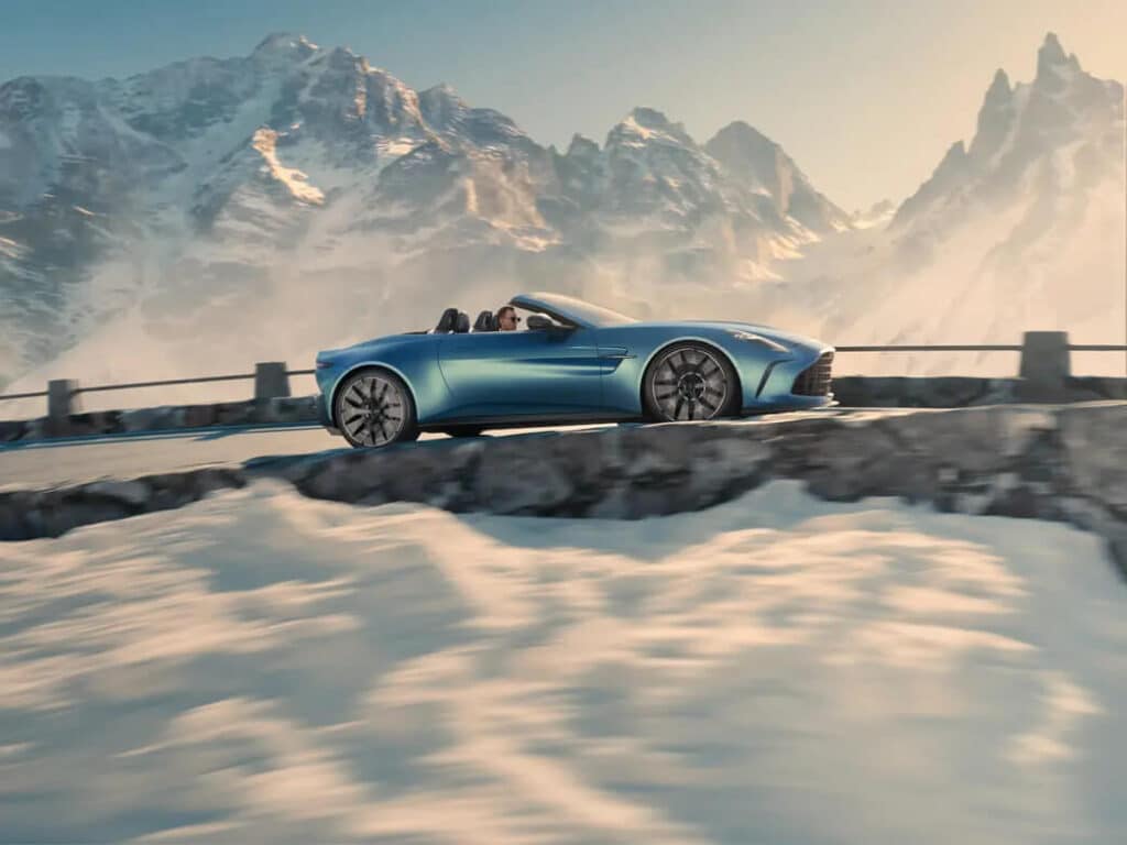 Imagen de un coche deportivo descapotable azul conduciendo por una carretera de montaña con picos nevados al fondo. El coche se desplaza por una carretera con una barrera de piedra al lado, y la escena se desarrolla en un paisaje majestuoso de montañas cubiertas de nieve bajo un cielo despejado. La imagen es interesante y relevante porque captura el contraste entre el diseño moderno y elegante del coche y la belleza natural y robusta del paisaje montañoso.