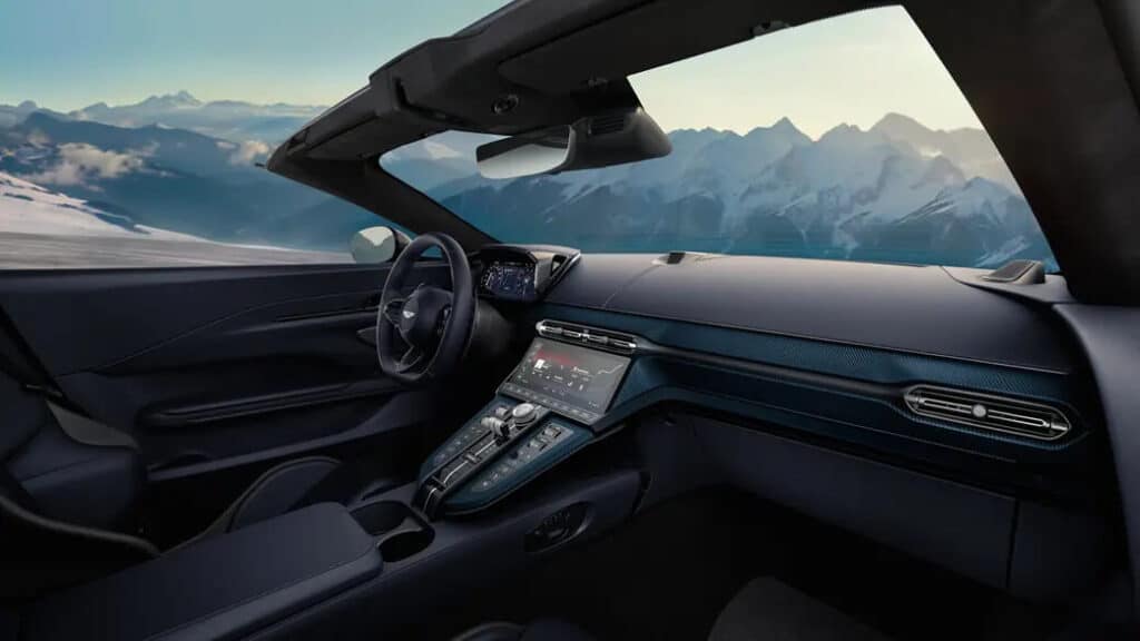 Imagen del interior de un Aston Martin Vantage Roadster 2025. El diseño interior es moderno y elegante, con un enfoque en el lujo y la tecnología. El salpicadero incluye una gran pantalla táctil con varios controles y configuraciones, un cuadro de instrumentos digital detrás del volante y materiales de alta calidad en todo el habitáculo. El techo descapotable está bajado, proporcionando una vista clara del paisaje montañoso en el fondo, lo que añade una sensación de aventura y lujo asociada con este vehículo.