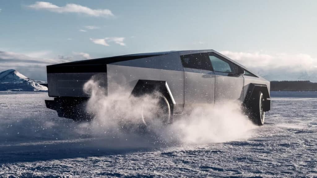 Vehículo futurista y angular conduciendo sobre un terreno nevado. El vehículo está levantando nieve mientras se mueve, lo que indica que está en movimiento. El fondo presenta una montaña cubierta de nieve y un cielo parcialmente nublado. Esta imagen destaca el diseño único y moderno de la Cybertruck y su rendimiento en condiciones de nieve.