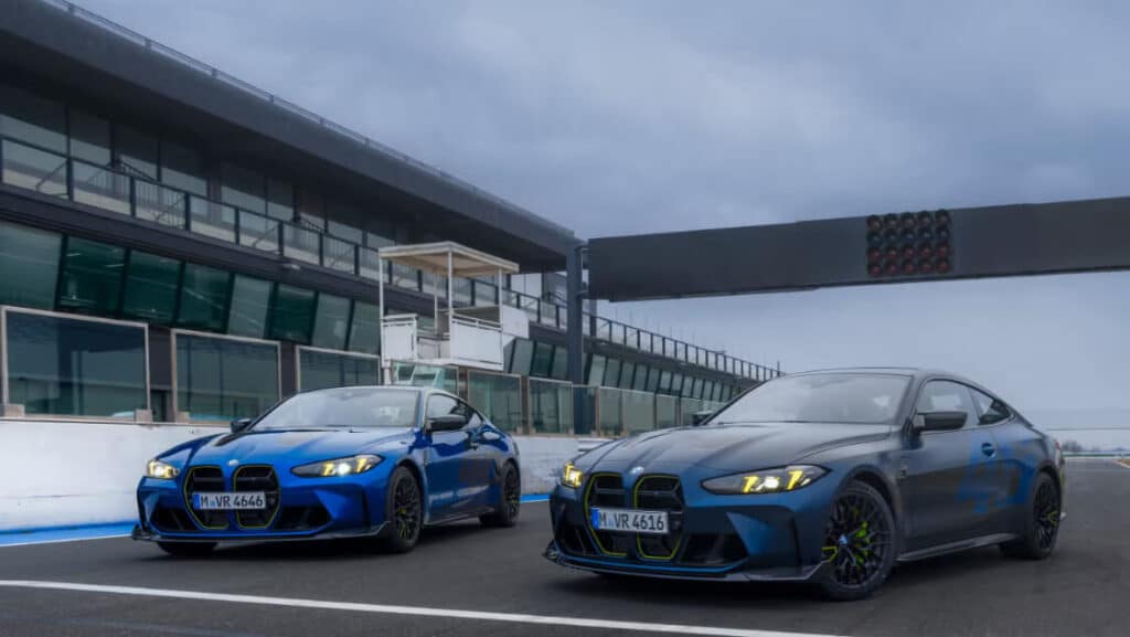 : Dos coches deportivos BMW M4 CS VR46 están estacionados en una pista de carreras. El coche a la izquierda es de color azul brillante con matrícula "M VR 4646", mientras que el coche a la derecha es de color azul oscuro con matrícula "M VR 4616". Ambos coches tienen los faros encendidos y están frente a una estructura de pista de carreras con luces de señalización en la parte superior.