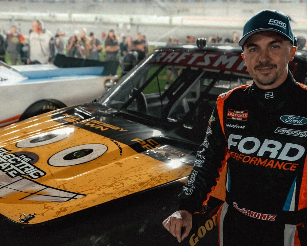 La imagen muestra a Frankie Muniz, conocido por su papel en la serie "Malcolm in the Middle", en un entorno de carreras. Lleva un traje de carreras con varios logos de patrocinadores, incluyendo "FORD PERFORMANCE" prominentemente visible en el pecho. El casco también tiene "FORD PERFORMANCE" escrito en él. El nombre "Frankie Muniz" es visible en la zona de la cintura del traje de carreras. El coche de carreras tiene un diseño distintivo en el capó, con una cara caricaturesca con grandes ojos y una gran sonrisa. En el fondo, hay varias personas y otro coche de carreras, lo que indica que esta imagen fue tomada en un evento de carreras. Esta imagen es interesante porque muestra a Frankie Muniz, un conocido actor, en un rol diferente como piloto de carreras, destacando su participación en el automovilismo.