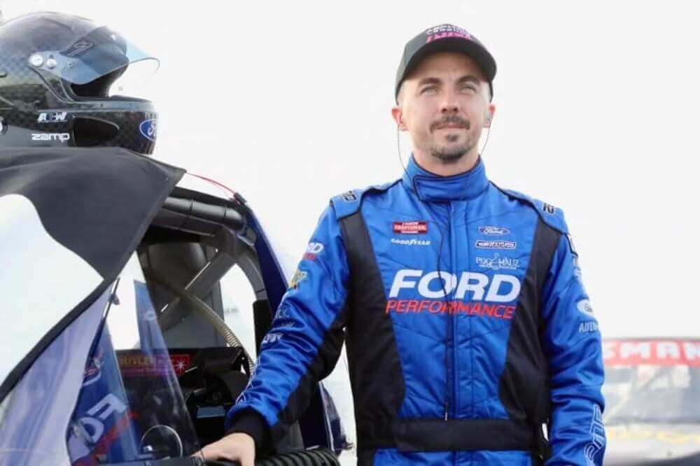 : La imagen muestra a Frankie Muniz, conocido por su papel en la serie "Malcolm in the Middle", vestido con un traje de carreras azul con la inscripción "FORD PERFORMANCE" en el frente. Está de pie junto a un coche de carreras, con un casco colocado sobre el coche. El traje de carreras también tiene varios logotipos de patrocinadores, incluyendo "Goodyear" y "Pro-Matic Automation". La imagen captura a Muniz en su papel como piloto de automovilismo, destacando su participación en la industria del deporte motor.