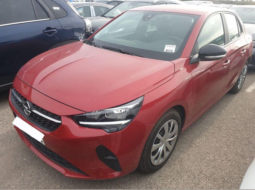 Una imagen de un Opel Corsa 2023 en Zaragoza estacionado en una calle de Zaragoza. El coche es de color rojo brillante y destaca con su diseño aerodinámico y moderno. En el fondo, se puede apreciar una vista urbana típica de Zaragoza, con edificios de varios pisos y árboles a lo largo de la calle. El Opel Corsa está en primer plano y ocupa la mayor parte de la imagen, mostrando su parte frontal y lateral.