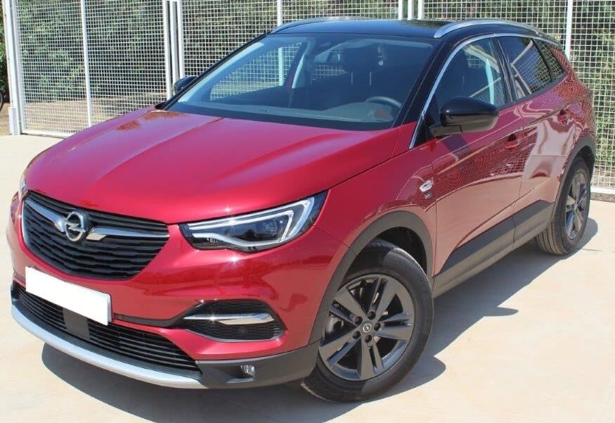 Un Opel Grandland X 2021 seminuevo de color rojo brillante está estacionado en un área pavimentada. El vehículo es un SUV con un diseño moderno y elegante. La parrilla frontal es negra con el logotipo de Opel en el centro. Los faros delanteros son de diseño angular y tienen luces LED. Las llantas son de aleación y de color oscuro. El coche tiene un techo de color negro que contrasta con el color rojo del cuerpo. No hay personas visibles en la imagen.