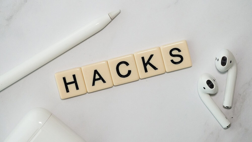 La imagen muestra un fondo blanco con varios objetos organizados sobre él. En la esquina superior izquierda hay un lápiz óptico blanco, en el centro hay cinco baldosas beige con letras negras que forman la palabra "HACKS", y a la derecha hay un par de auriculares inalámbricos blancos. Además, hay una funda de carga blanca para los auriculares en la parte inferior izquierda. Esta imagen combina elementos relacionados con la tecnología (lápiz óptico y auriculares inalámbricos) con la palabra "HACKS", lo que sugiere consejos o trucos relacionados con la tecnología.