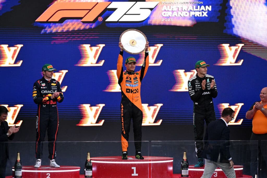 Ceremonia del podio del Gran Premio de Australia de Fórmula 1 2025. El piloto que quedó en primer lugar, vistiendo un mono de carreras naranja de McLaren, sostiene un trofeo sobre su cabeza. A su izquierda está el piloto en segundo lugar, con un mono de Red Bull, y a su derecha el tercero, con un mono de Mercedes. El fondo muestra el logo de Louis Vuitton y el nombre del evento. Hay botellas de champán Moët en los escalones del podio.