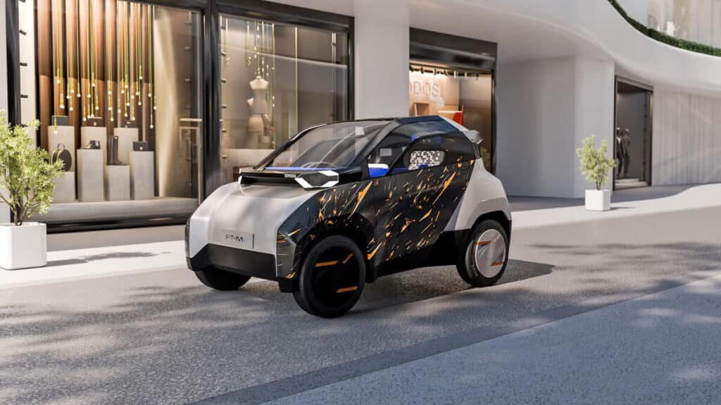 Imagen de un coche compacto futurista, el Toyota FT-M, estacionado frente a una moderna tienda con grandes ventanales. El coche presenta un diseño elegante en colores blanco y negro, con detalles en naranja y un patrón decorativo que recuerda a chispas en el lateral. La escena está iluminada con luz diurna, destacando el entorno urbano contemporáneo.
