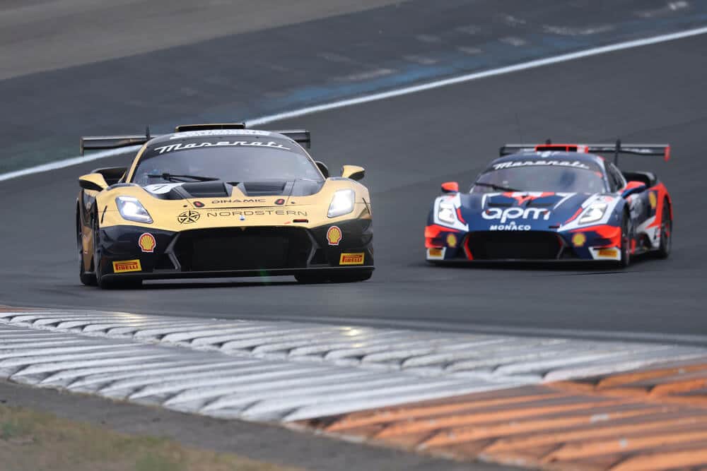 "Dos coches de carreras Maserati avanzan a gran velocidad sobre la pista. El coche de la izquierda tiene un diseño dorado con detalles negros y luce los logotipos de Maserati, Dinamic, Nordstern, Shell y Pirelli. Su alerón trasero grande y su carrocería aerodinámica resaltan su enfoque en el rendimiento. A la derecha, otro Maserati, con una base negra y detalles en rojo y blanco, lleva los logotipos de Maserati, apm Monaco, Shell y Pirelli. Ambos autos están en plena competencia, con el coche dorado ligeramente adelantado, capturando la esencia de la velocidad y la emoción del automovilismo.