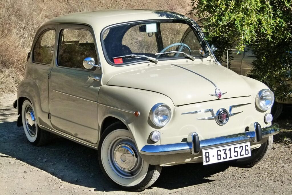 Un Seat 600 clásico, de color beige, está aparcado sobre un suelo de tierra. El coche presenta un diseño icónico con líneas redondeadas y detalles cromados. En la parte frontal destacan su parrilla con el logotipo de Seat, faros redondos y una matrícula con el número 'B-631526'. En el fondo, se pueden ver arbustos y vegetación, lo que sugiere un entorno al aire libre. Este modelo fue un símbolo de movilidad en España durante el siglo XX, apreciado por su tamaño compacto y fiabilidad.